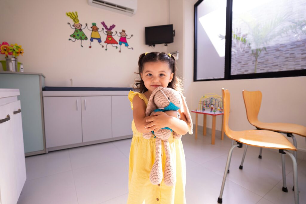 A cheerful girl in a yellow dress hugging a stuffed toy in a bright room with playful decor.